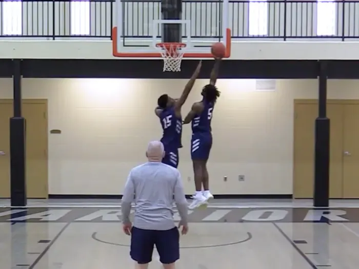 Players running a layup drill at the basket with a coach watching from mid-court